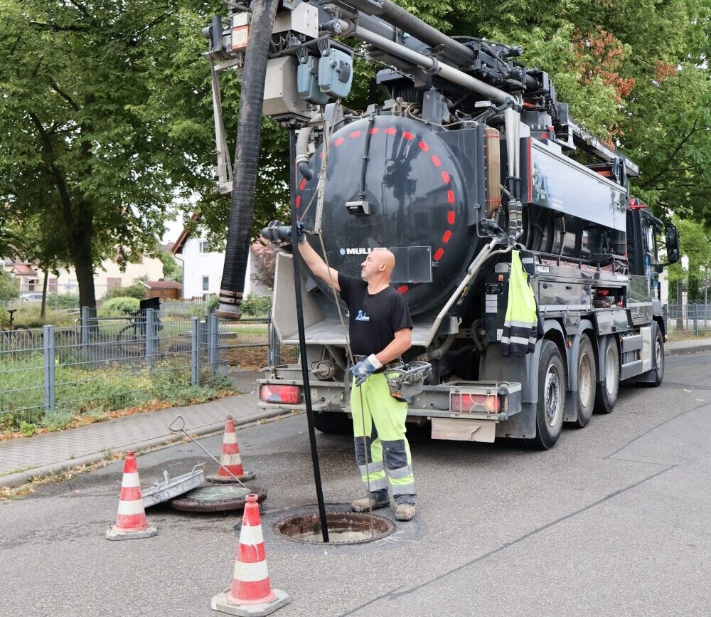 Ein Mitarbeiter des Kanalserviceteams arbeitet mit einem Saug- und Spülwagen an einem geöffneten Kanalschacht. Er führt einen Schlauch in den Schacht ein, während mehrere Warnkegel die Arbeitsstelle absichern.